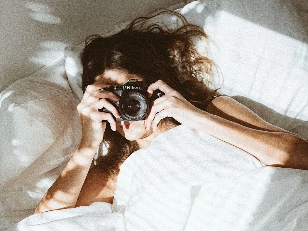 A person lying in bed under white sheets holds a Nikon camera up to their face, preparing to take a photo. Sunlight and shadows fall across the scene.
