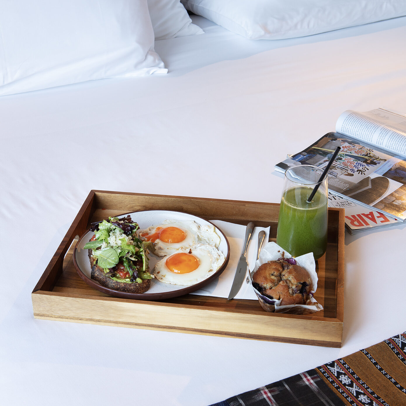 Breakfast tray with avocado toast, fried eggs, a muffin, and green juice on a white hotel bed.